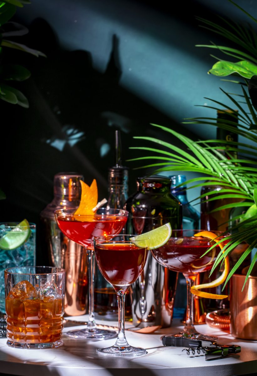 Summer alcoholic cocktail party with ready-made cocktails, bar tools and bottles of spirits on marble table with tropical plants. Green background, hard light and shadows. Shallow DOF